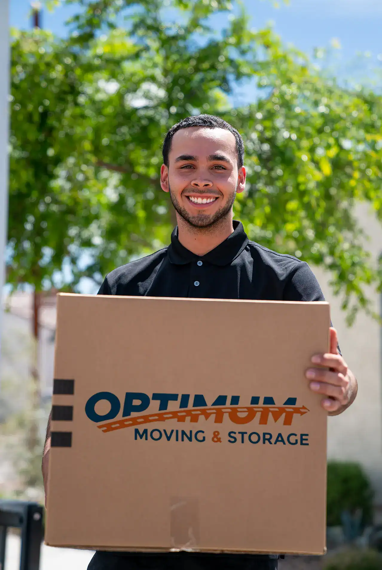 Professional mover holding a box with Optimum Moving & Storage logo outdoors.